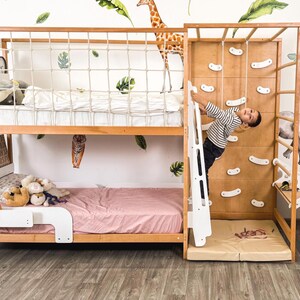 Pu&ograve; includere: Un letto a castello in legno con una parete da arrampicata e una scala. Il letto ha una struttura marrone chiaro con una rete di sicurezza bianca sulla cuccetta superiore. Un bambino sta arrampicando sul muro. La stanza &egrave; decorata con adesivi murali a forma di foglie e animali.