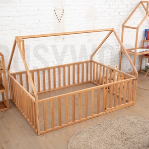 May include: A wooden playpen with a house-shaped frame, featuring vertical slats and a small door. The playpen is made of light-colored wood and is surrounded by other wooden furniture, including a bookshelf and a desk. A beige rug is on the floor.