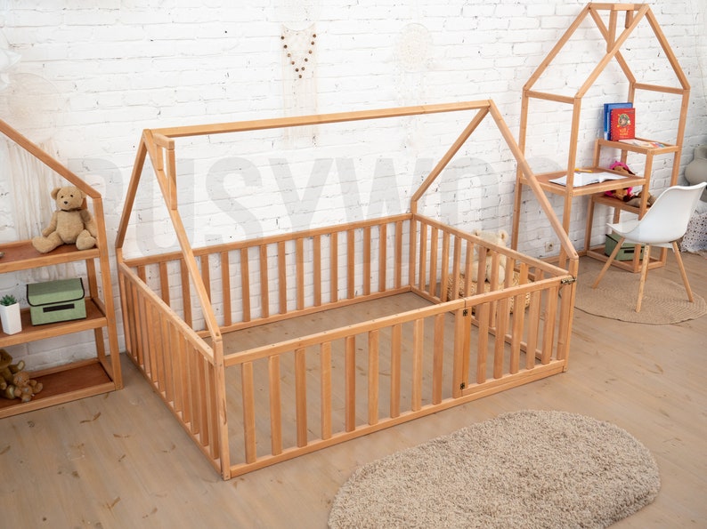 May include: A wooden playpen with a house-shaped frame, featuring vertical slats and a small door. The playpen is made of light-colored wood and is surrounded by other wooden furniture, including a bookshelf and a desk. A beige rug is on the floor.