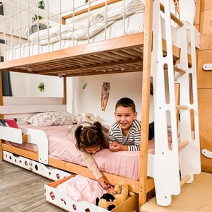 Pu&ograve; includere: Un letto a castello in legno con una scala bianca e una rete di corda sulla cuccetta superiore. La cuccetta inferiore ha un lenzuolo rosa e due bambini che giocano. Un cassetto estraibile &egrave; aperto, rivelando giocattoli.