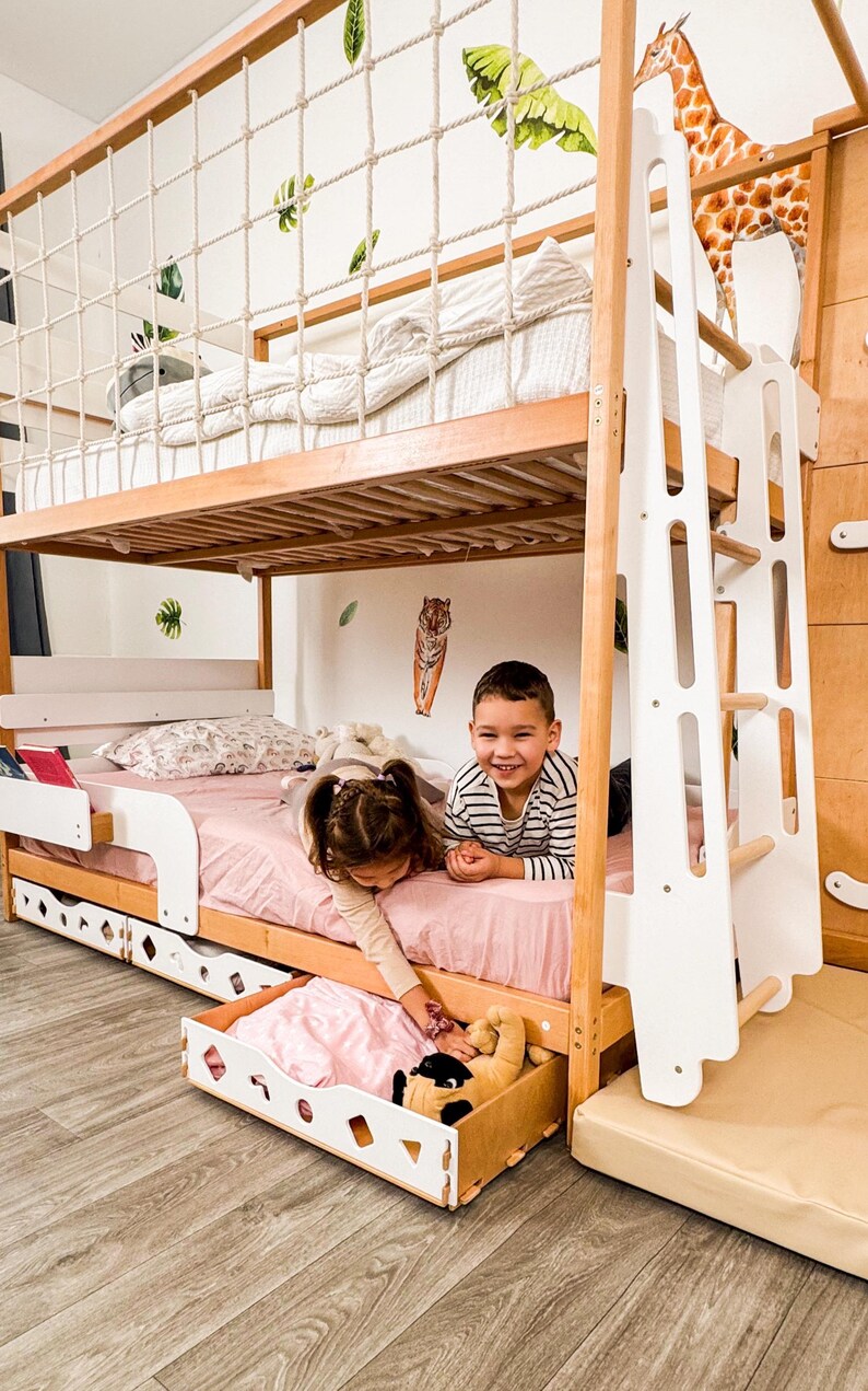 Pu&ograve; includere: Un letto a castello in legno con una scala bianca e una rete di corda sulla cuccetta superiore. La cuccetta inferiore ha un lenzuolo rosa e due bambini che giocano. Un cassetto estraibile &egrave; aperto, rivelando giocattoli.