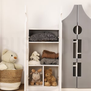 Op de afbeelding: Witte houten kast met twee deuren en planken. De kast is open en gevuld met knuffels en dekens. De kast staat in een kamer met een houten vloer.