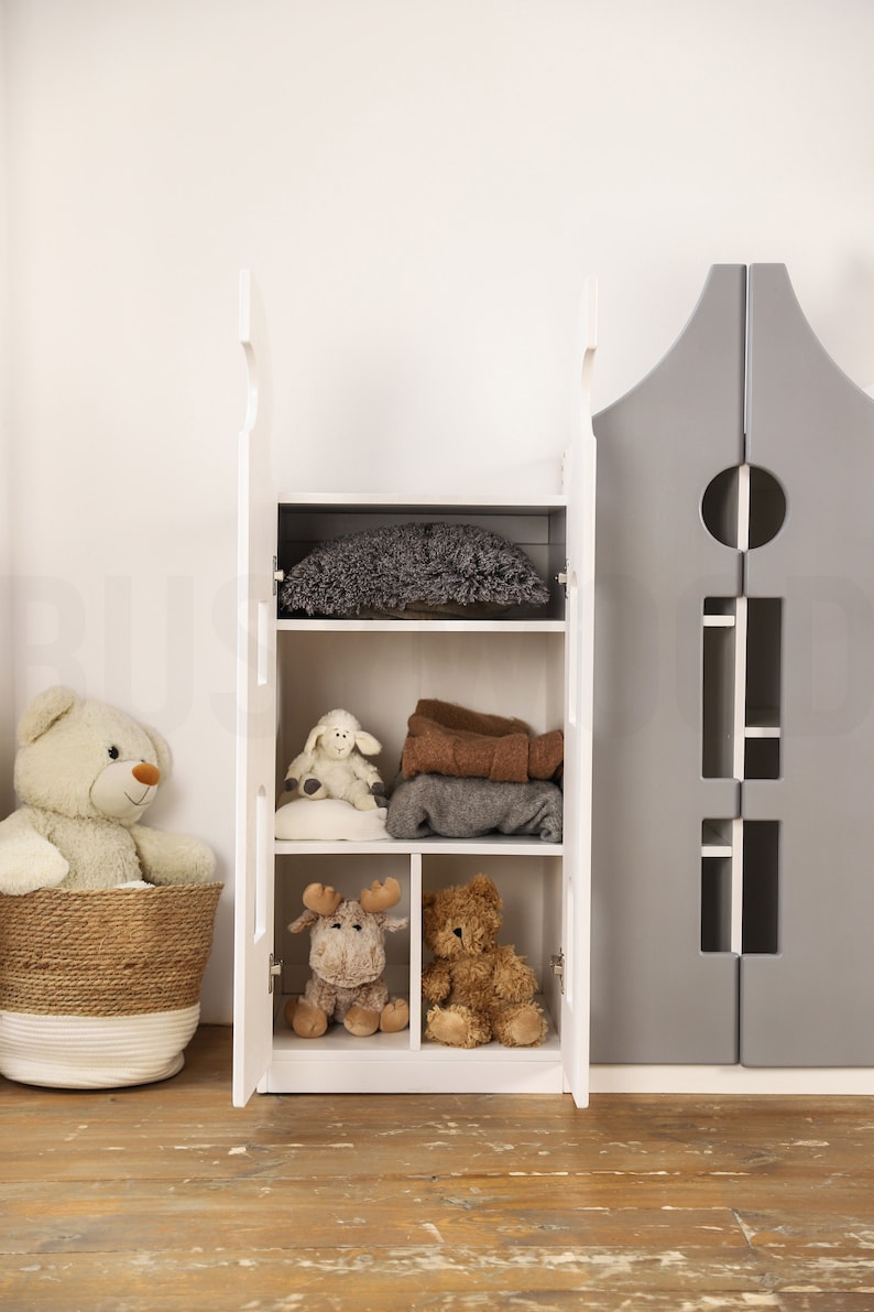 Op de afbeelding: Witte houten kast met twee deuren en planken. De kast is open en gevuld met knuffels en dekens. De kast staat in een kamer met een houten vloer.