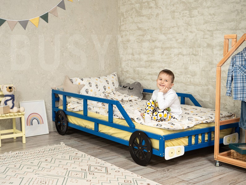 K&ouml;nnte beinhalten: Ein blaues Holzbettgestell mit schwarzen R&auml;dern, einer gelben Matratze und einem wei&szlig;en und gelben Bettw&auml;sche-Set. Ein kleiner Junge sitzt auf dem Bett, l&auml;chelt und schaut in die Kamera.
