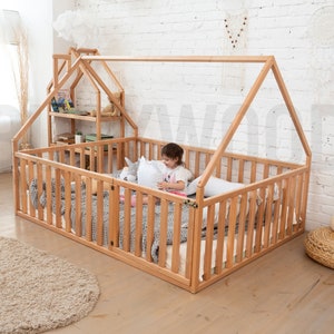 May include: A natural wood house-shaped bed frame with a slatted side and a matching small house-shaped shelf unit. The bed is filled with pillows and blankets. A child is sitting inside the bed. A beige rug and a woven ottoman are in the foreground.