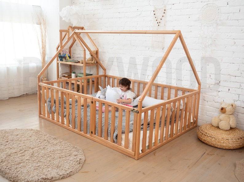 May include: A natural wood house-shaped bed frame with a slatted side and a matching small house-shaped shelf unit. The bed is filled with pillows and blankets. A child is sitting inside the bed. A beige rug and a woven ottoman are in the foreground.
