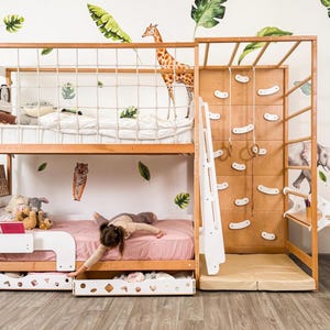 Pu&ograve; includere: Un letto a castello in legno con una parete da arrampicata, una scala e una rete di corda. Il letto ha una struttura in legno chiaro con un materasso rosa sulla cuccetta inferiore. Un bambino sta giocando sul letto. Sono visibili adesivi murali a tema giungla.