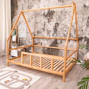 May include: A wooden teepee-style bed frame with a slatted mattress and a safety rail. The bed is in a room with a light brown wood floor and a textured gray wall.