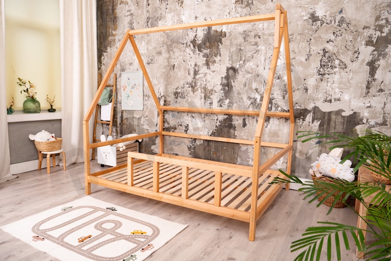 May include: A wooden teepee-style bed frame with a slatted mattress and a safety rail. The bed is in a room with a light brown wood floor and a textured gray wall.