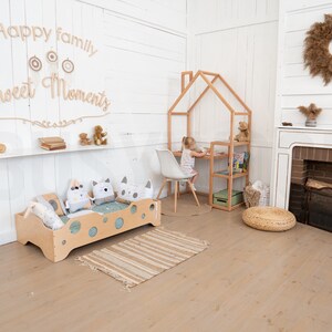 May include: A light wood children's bed with a white and gray patterned pillow and a light wood playhouse with a shelf and a small table and chair. The text "Happy family Sweet Moments" is on the wall behind the bed.