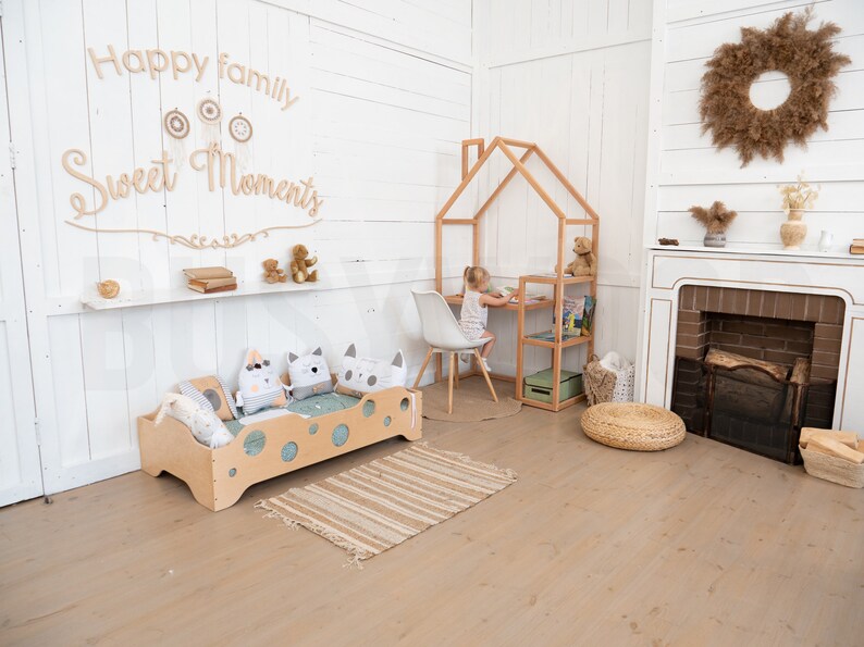 May include: A light wood children's bed with a white and gray patterned pillow and a light wood playhouse with a shelf and a small table and chair. The text "Happy family Sweet Moments" is on the wall behind the bed.