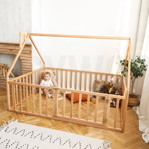 May include: A natural wood house-shaped playpen with a gated door. Inside, a child sits with a teddy bear. The playpen has a slatted design and is set on a wooden floor. A small rug is in the foreground.