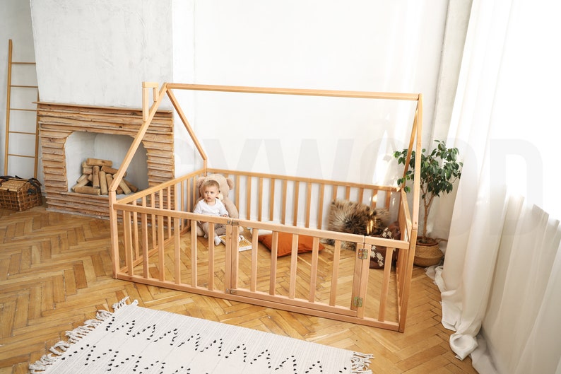 May include: A natural wood house-shaped playpen with a gated door. Inside, a child sits with a teddy bear. The playpen has a slatted design and is set on a wooden floor. A small rug is in the foreground.