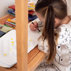 K&ouml;nnte beinhalten: Ein junges M&auml;dchen sitzt an einem Tisch und malt mit einem schwarzen Stift in einem Buch. Sie tr&auml;gt ein wei&szlig;es Hemd mit einem schwarz-wei&szlig;en Muster aus kleinen Tieren. Hinter ihr liegen mehrere B&uuml;cher auf dem Tisch.