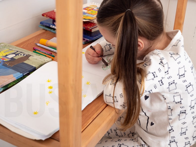 K&ouml;nnte beinhalten: Ein junges M&auml;dchen sitzt an einem Tisch und malt mit einem schwarzen Stift in einem Buch. Sie tr&auml;gt ein wei&szlig;es Hemd mit einem schwarz-wei&szlig;en Muster aus kleinen Tieren. Hinter ihr liegen mehrere B&uuml;cher auf dem Tisch.