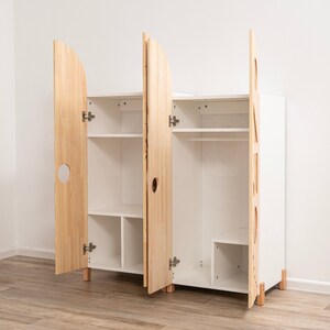 May include: A white wooden wardrobe with two doors, one with a round cutout and the other with a geometric cutout. The doors are open, revealing shelves and a hanging rod inside.