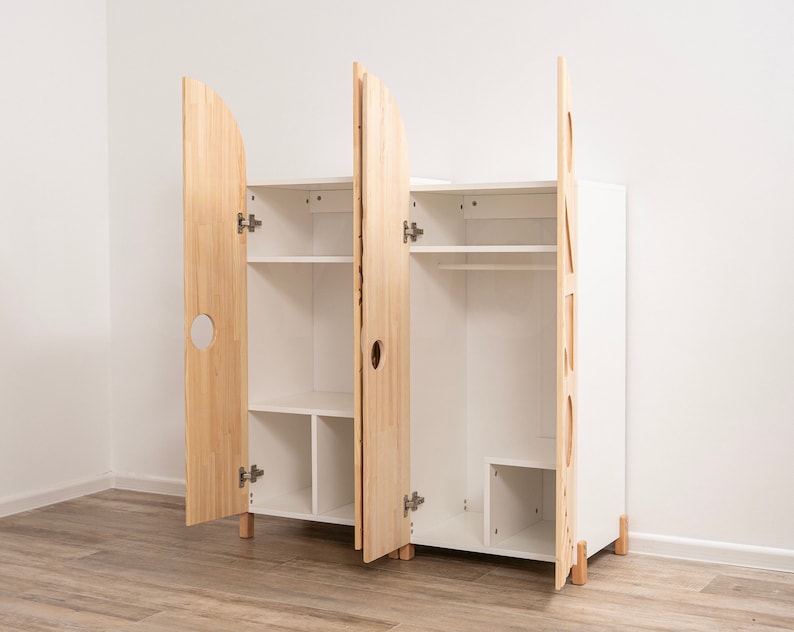 May include: A white wooden wardrobe with two doors, one with a round cutout and the other with a geometric cutout. The doors are open, revealing shelves and a hanging rod inside.