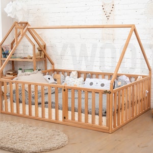 May include: A natural wood-framed toddler bed with a house-shaped frame. The bed has a slatted side and a small shelf unit. Pillows and a blanket are inside the bed. A beige rug is in front of the bed.