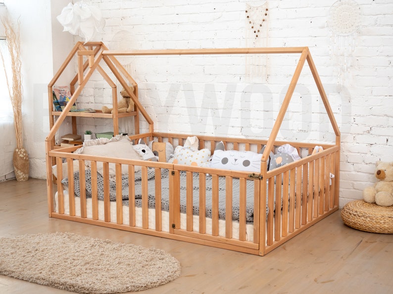 May include: A natural wood-framed toddler bed with a house-shaped frame. The bed has a slatted side and a small shelf unit. Pillows and a blanket are inside the bed. A beige rug is in front of the bed.
