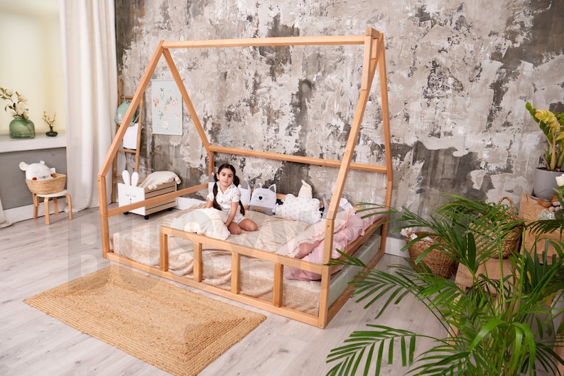 May include: A natural wood-framed bed in a house shape, with a mattress and pillows. A young person sits on the bed. The room has a neutral color palette, with a jute rug and decorative items.