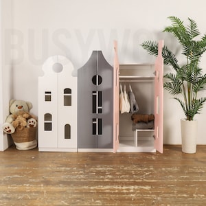 Op de afbeelding: Drie speelgoedhuisvormige opbergkasten met deuren. De kast links is wit, de middelste kast is grijs en de kast rechts is roze. De roze kast heeft een hangstang en planken erin.