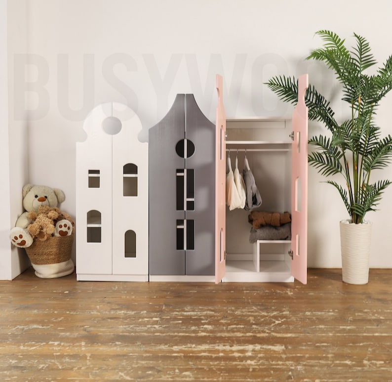 Op de afbeelding: Drie speelgoedhuisvormige opbergkasten met deuren. De kast links is wit, de middelste kast is grijs en de kast rechts is roze. De roze kast heeft een hangstang en planken erin.