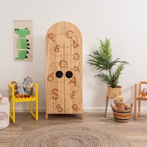 May include: A wooden children's wardrobe with a whimsical design featuring the alphabet in a continuous line. The wardrobe has two doors and is painted in a natural wood finish. There are two small round holes on the doors. The wardrobe is surrounded by a variety of toys and decor, including stuffed animals, a yellow chair, and a potted plant.