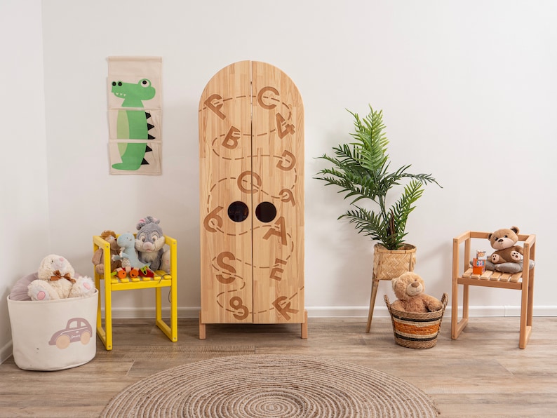 May include: A wooden children's wardrobe with a whimsical design featuring the alphabet in a continuous line. The wardrobe has two doors and is painted in a natural wood finish. There are two small round holes on the doors. The wardrobe is surrounded by a variety of toys and decor, including stuffed animals, a yellow chair, and a potted plant.