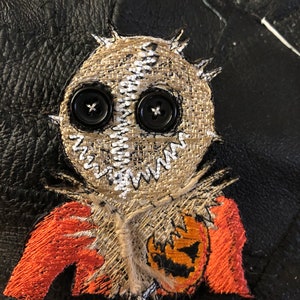 May include: Embroidered patch of a burlap scarecrow with black button eyes and a stitched mouth. The scarecrow is wearing a red and orange shirt with a jack-o-lantern design.