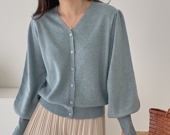 May include: A light blue cardigan with a V-neck and button closure. The cardigan has long sleeves with a puffed design. Paired with pleated beige pants. A woven basket sits on a wooden table.