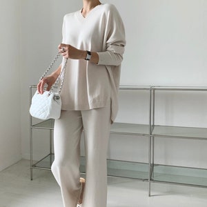 Könnte beinhalten: Beige Strick-Set bestehend aus einem V-Ausschnitt-Pullover und einer Hose mit weitem Bein. Das Outfit wird mit weißen Sandalen und einer gesteppten weißen Handtasche mit silberner Kette kombiniert. Das Ensemble ist schlicht und elegant.