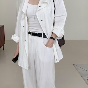 以下が含まれることがあります： 白いジャケット、タンクトップ、ワイドパンツのコーディネート。 ジャケットは襟とスナップ留め。 黒いベルトがウエストをマーク。 シルバーのネックレスと黒い腕時計が見える。 茶色のショルダーバッグがかけられている。