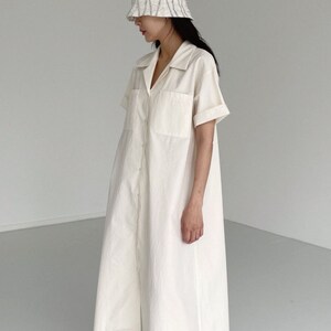 May include: A cream-colored, button-down dress with short sleeves and two front pockets. The dress is paired with a white and gray striped bucket hat and white flip-flops. The dress has a collared neckline and a relaxed fit.