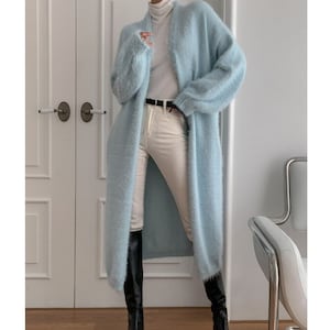 May include: A long, light blue cardigan with a fluffy texture. The cardigan is worn over a white turtleneck and cream-colored pants. Black knee-high boots complete the outfit. The cardigan is open in the front, revealing the outfit underneath.