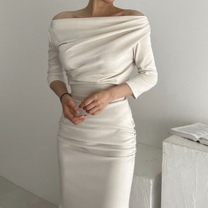 Peut inclure: Robe crème à épaules dénudées et manches longues. La robe présente un tissu drapé sur le corsage et des fronces sur la jupe. Le modèle porte un bracelet noir. Un livre est posé sur une surface blanche.