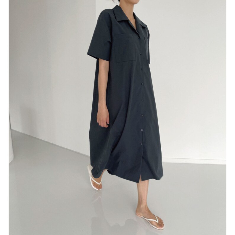 May include: A dark navy blue button-down shirt dress with short sleeves and a collared neckline. The dress features two chest pockets and a button-up front. The hemline falls below the knees. The person is wearing white flip-flops.