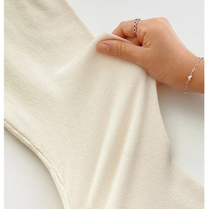 May include: Close-up of a cream-colored fabric being held. The fabric appears soft and slightly textured. A hand with a silver ring and bracelet is holding the fabric, showing its flexibility and drape.