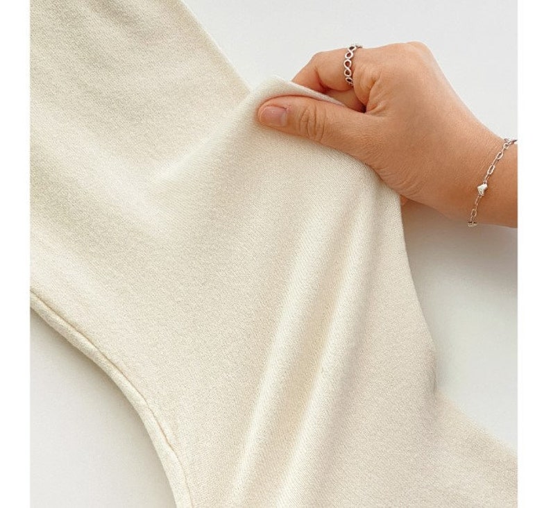 May include: Close-up of a cream-colored fabric being held. The fabric appears soft and slightly textured. A hand with a silver ring and bracelet is holding the fabric, showing its flexibility and drape.