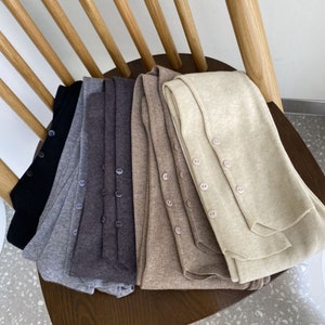 Peut inclure: Plusieurs cardigans boutonn&eacute;s de couleurs neutres, dont noir, gris et beige, sont pr&eacute;sent&eacute;s sur une chaise en bois. Chaque cardigan est dot&eacute; d'une rang&eacute;e de boutons et d'une texture c&ocirc;tel&eacute;e.