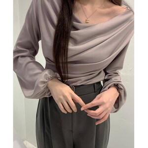 May include: A draped, mauve-colored blouse with long sleeves and a gathered neckline. The blouse is paired with dark gray trousers. A gold necklace is visible.