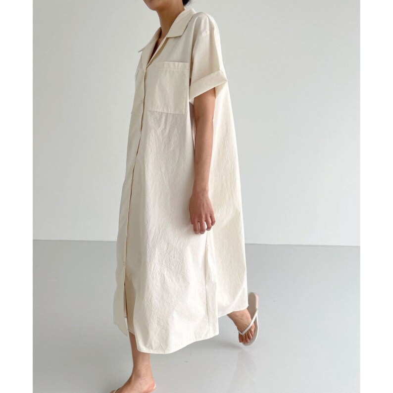 May include: A cream-colored, button-down dress with short sleeves and two front pockets. The dress has a collared neckline and a relaxed fit, reaching to the mid-calf. The model is wearing white flip-flops.