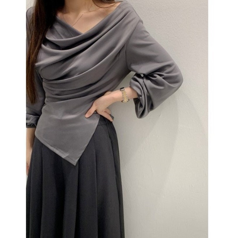 May include: A gray draped top with long sleeves and a cowl neckline paired with a black skirt. The top has an asymmetrical hemline. The outfit is simple and elegant, suitable for various occasions.