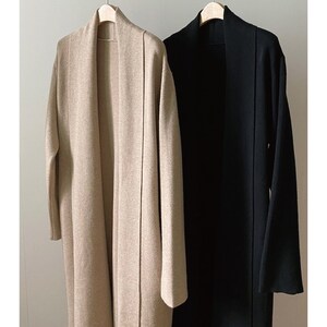 May include: Two long, open-front cardigans hanging on wooden hangers. One is a light beige color, and the other is solid black. The cardigans have a simple, minimalist design, with long sleeves and a flowing silhouette. The fabric appears to be a soft, medium-weight material.