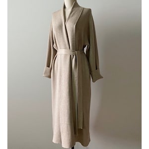 May include: A long, beige robe with a shawl collar and a matching belt. The robe has long sleeves with folded cuffs and is displayed on a mannequin. The fabric appears to be a soft, woven material.