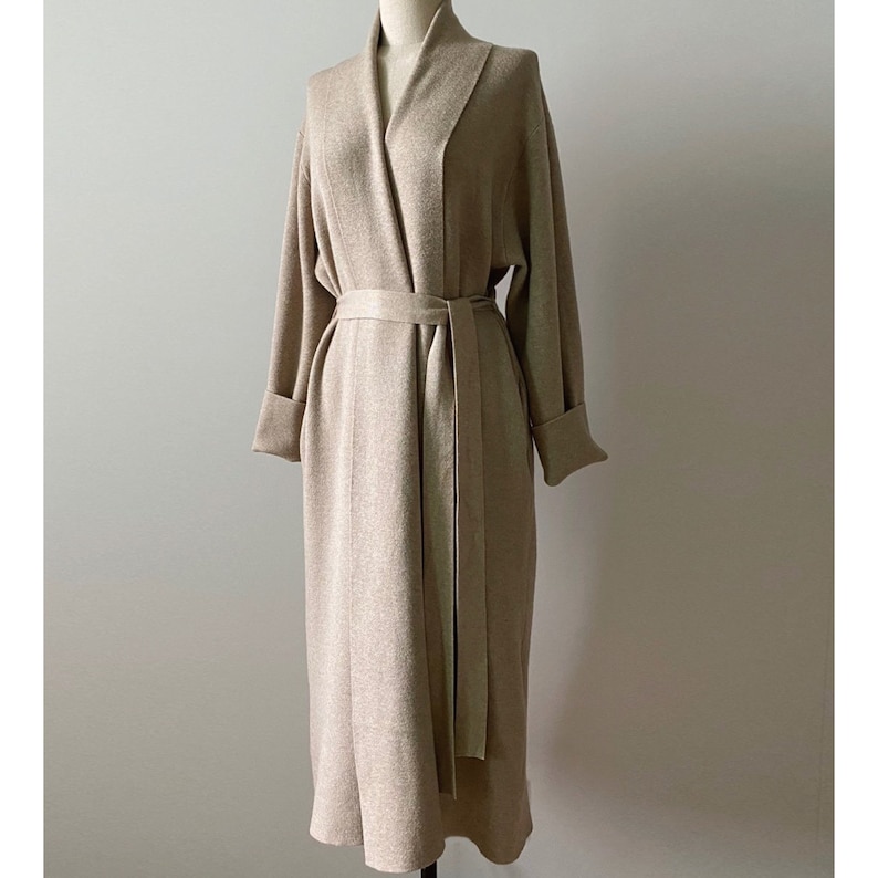 May include: A long, beige robe with a shawl collar and a matching belt. The robe has long sleeves with folded cuffs and is displayed on a mannequin. The fabric appears to be a soft, woven material.