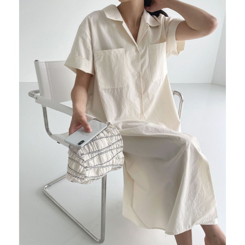 May include: A cream-colored, button-down shirt dress with short sleeves and two front pockets. The dress has a collared neckline and is worn with a small, white, ruched handbag. The person is seated on a white chair.