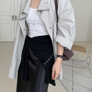 May include: A light grey trench coat with a white tank top and a black skirt. The coat has a button closure and a collar. The skirt has a wrap design. A brown shoulder bag is also visible.