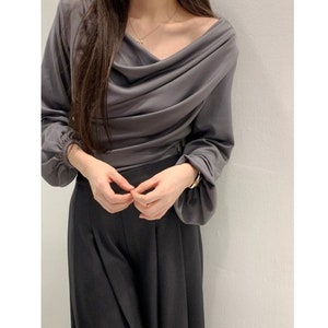 May include: A draped, long-sleeved gray top with a cowl neckline. The top is paired with dark gray, wide-leg pants. A gold necklace and bracelet are visible. The outfit is simple and elegant.