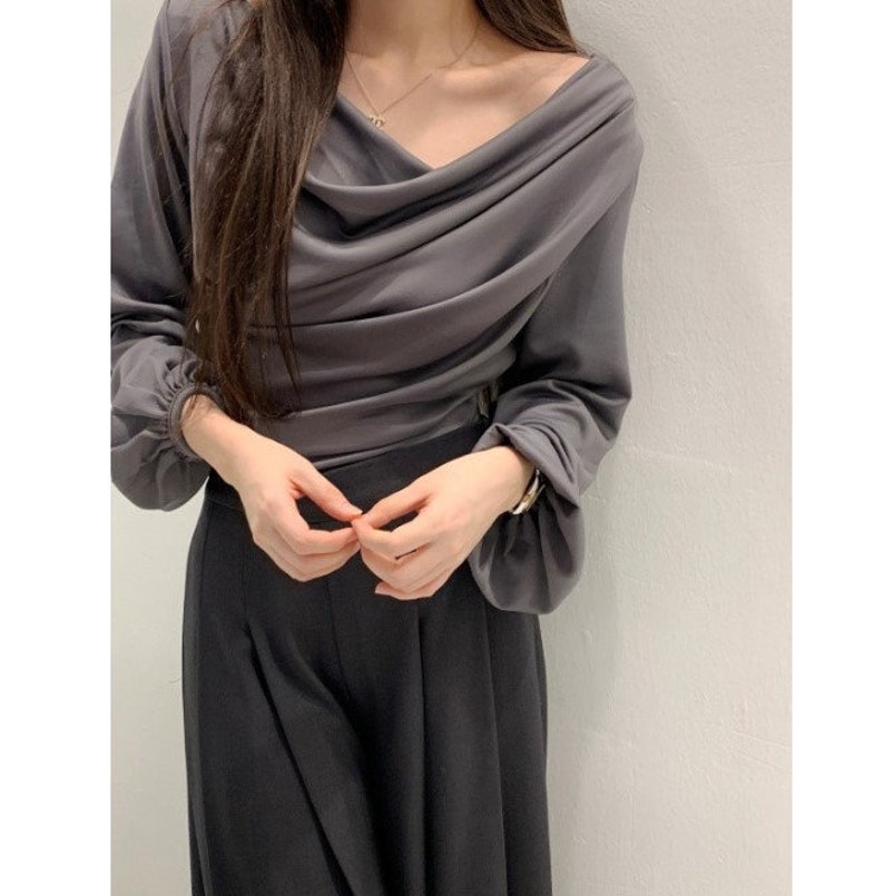 May include: A draped, long-sleeved gray top with a cowl neckline. The top is paired with dark gray, wide-leg pants. A gold necklace and bracelet are visible. The outfit is simple and elegant.