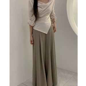 May include: A cream-colored long-sleeve top with an asymmetrical design and a draped neckline. Paired with wide-leg, pleated, olive-green pants. The outfit is a modern take on a classic style.